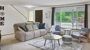 A modern living room with a sofa, armchairs, a coffee table, and a view of a garden through large windows. - Entire home in Port Orchard, Washington with Game Room, Fire Pit and RV Parking (Port Orchard)