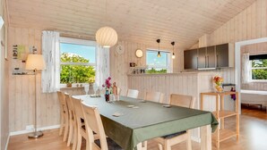 A spacious dining area with a long table, wooden chairs, and a green tablecloth. There is a large window providing natural light and a view of greenery outside.