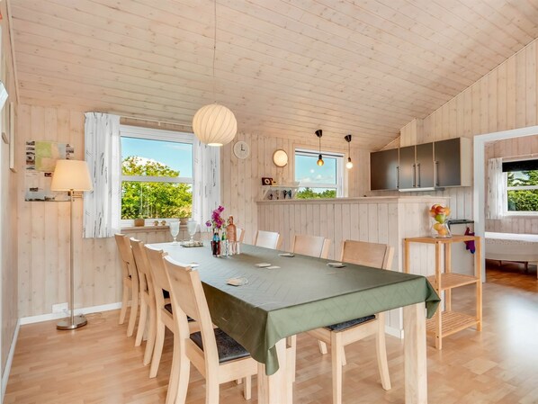 A spacious dining area with a long table, wooden chairs, and a green tablecloth. There is a large window providing natural light and a view of greenery outside.