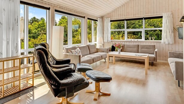 A spacious room with a wooden ceiling, large windows, a black leather chair, a grey sofa, a wooden coffee table, and a small table with a lamp.