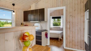 A compact kitchen with wooden walls, a dining area with a fruit bowl, and a bed in the adjacent room.
