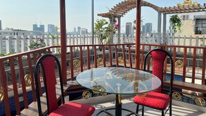 Un espacio de estar al aire libre techado con una mesa de vidrio y sillas rojas, con vistas a un paisaje urbano que incluye edificios y un cielo despejado.