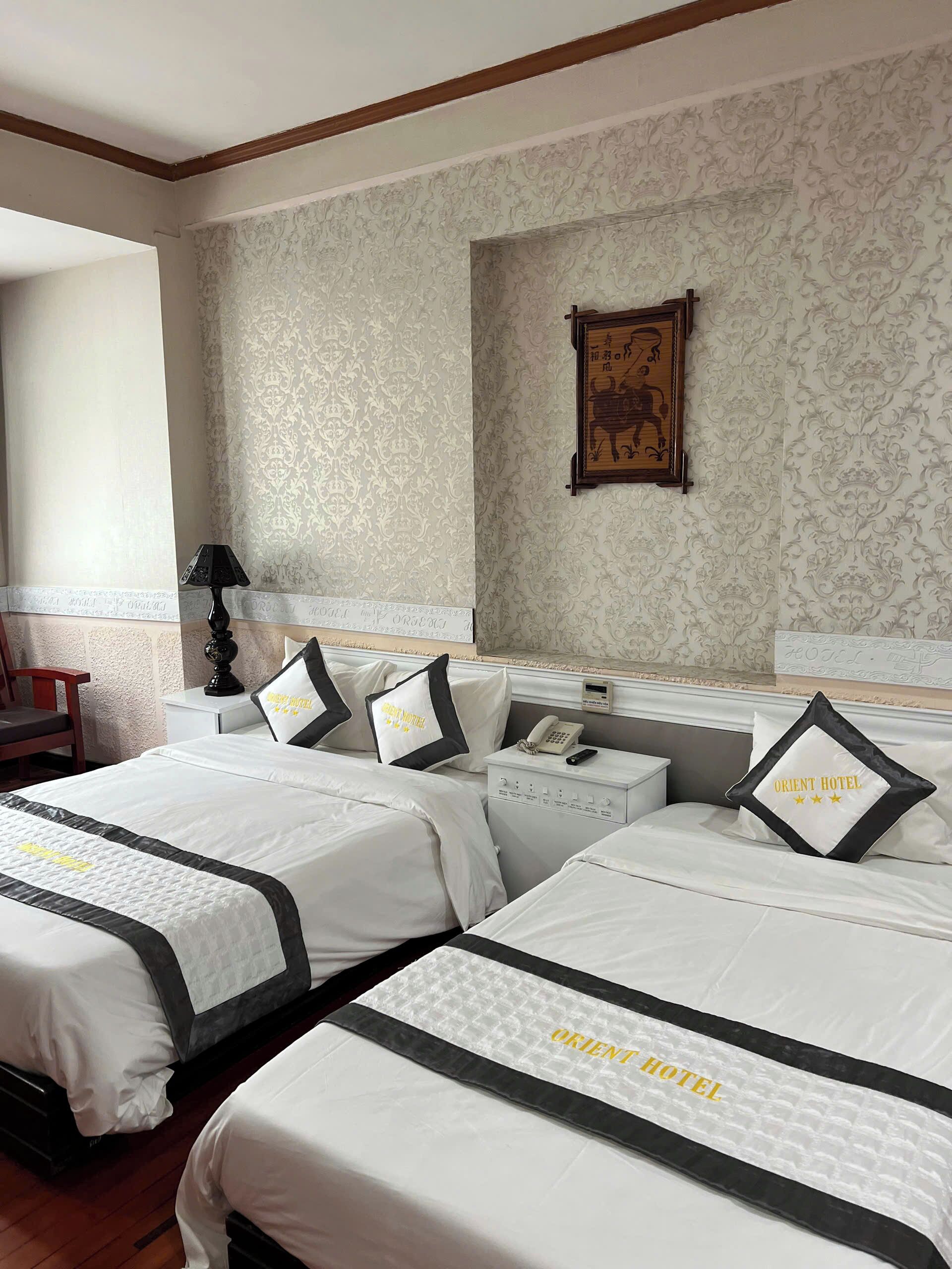 Habitación de hotel con dos camas, cada una con un almohadón que dice "ORIENT HOTEL". Hay una mesita de noche con un teléfono y una lámpara, y un cuadro en la pared.