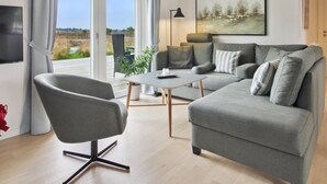 Un salon moderne avec un canapé gris, un fauteuil gris, une petite table et un lampadaire. On y trouve une fenêtre avec des rideaux, un tableau au mur et une vue sur l’extérieur.