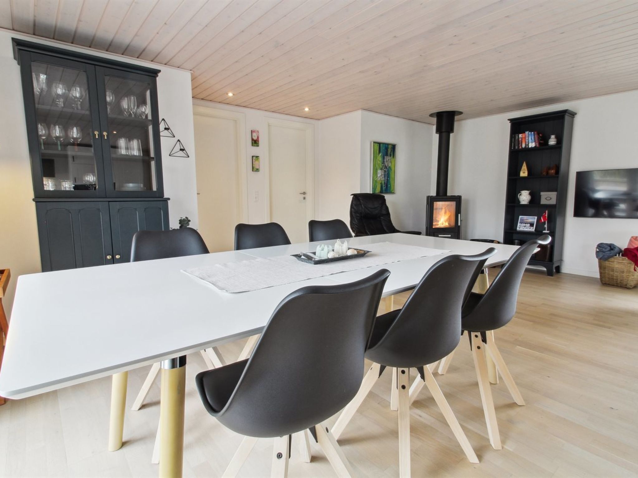 En moderne spisestue med et hvidt bord, sorte stole, et skab med glaslåger, en sort bogreol og en brændeovn.