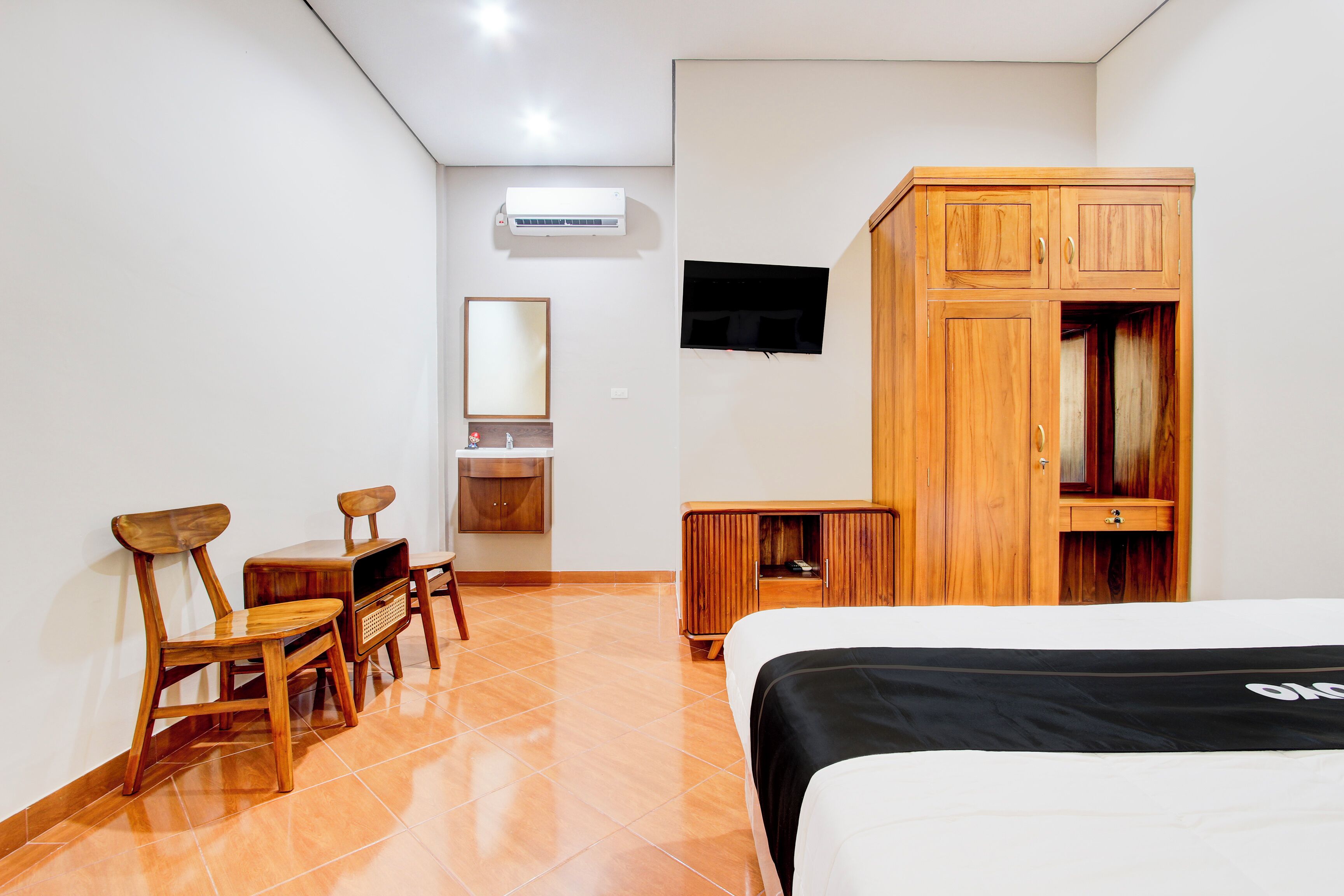 A hotel room with a bed, wooden wardrobe, TV mounted on the wall, and a desk with chairs.