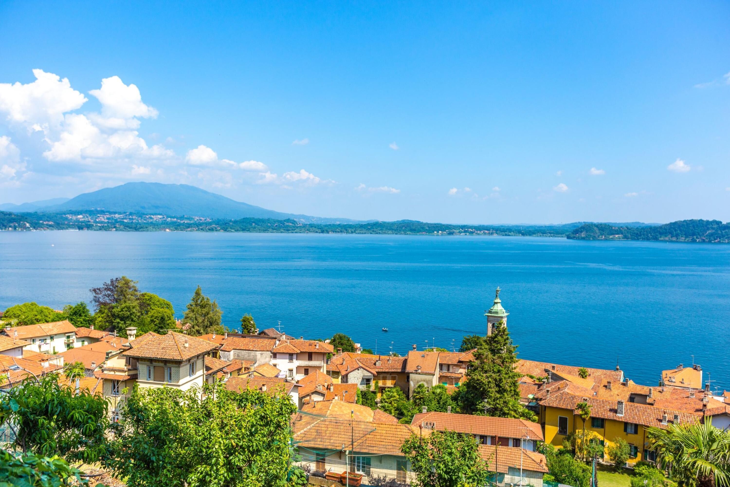 A coastal town with terracotta-roofed buildings, a clear blue sea, and a mountain in the background.