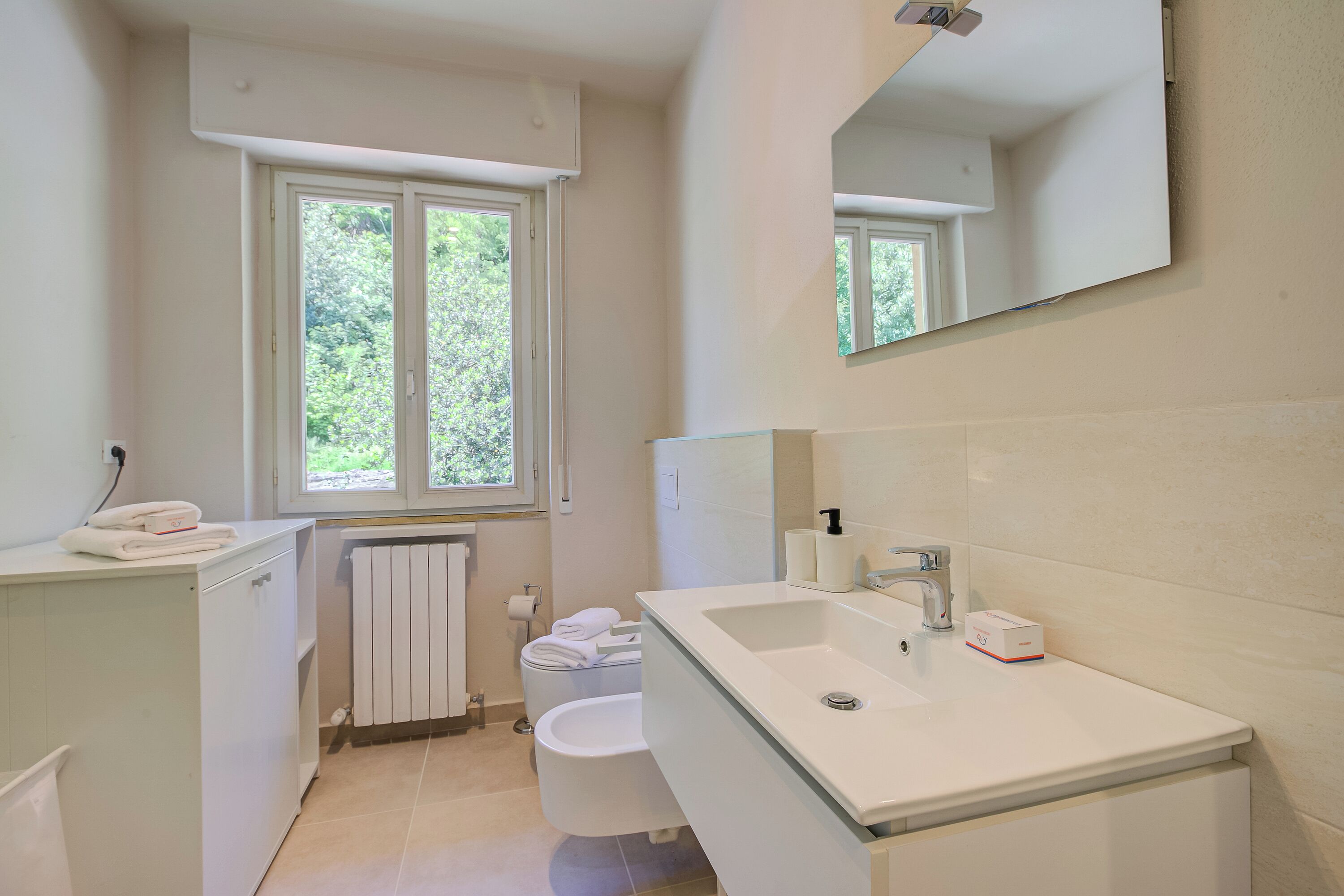 Un baño moderno con un lavamanos grande, un inodoro y un bidé. Hay una ventana que proporciona luz natural y un espejo sobre el lavamanos.