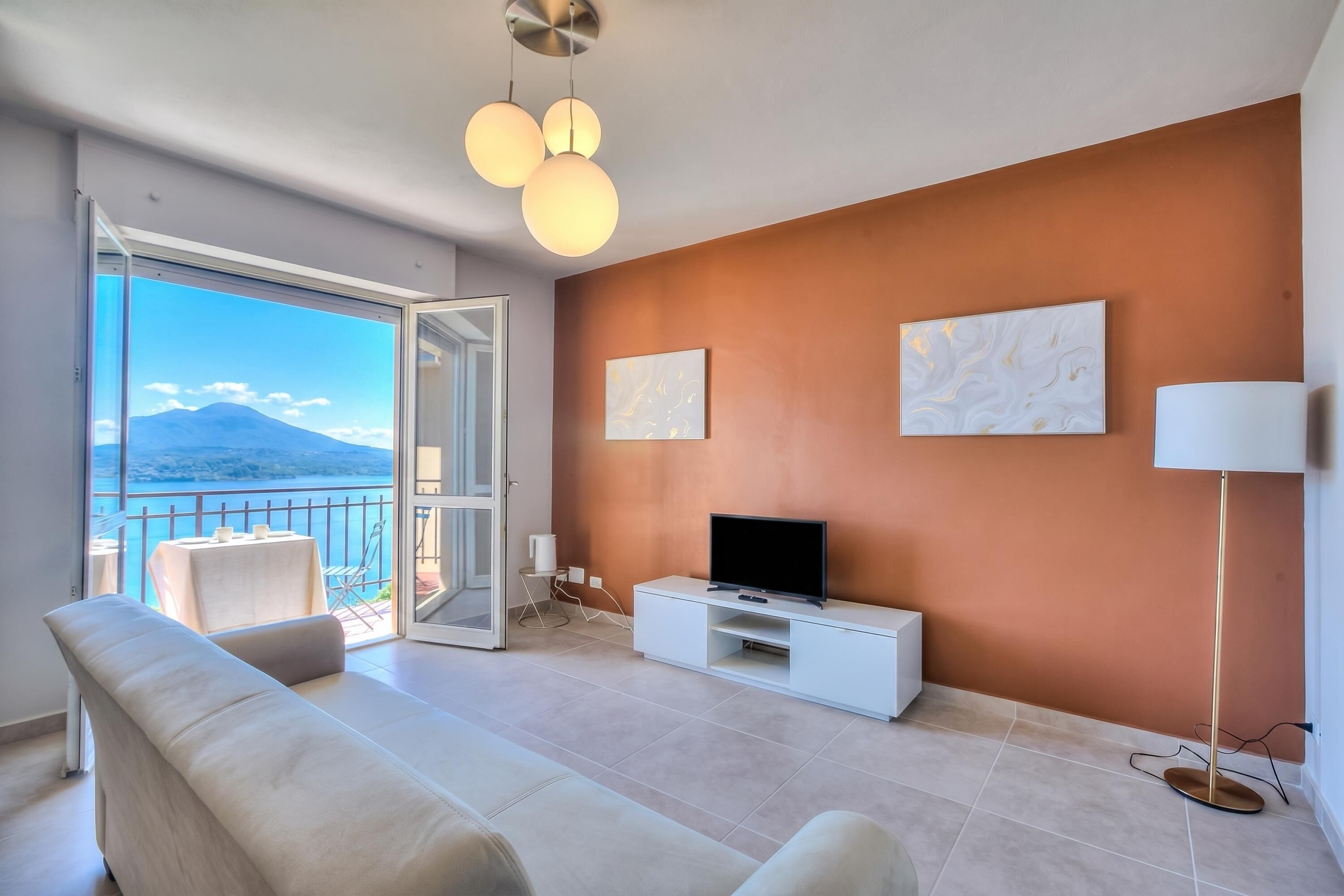A modern living room with a sofa, a TV on a white cabinet, a floor lamp, and a view of the sea through a balcony.