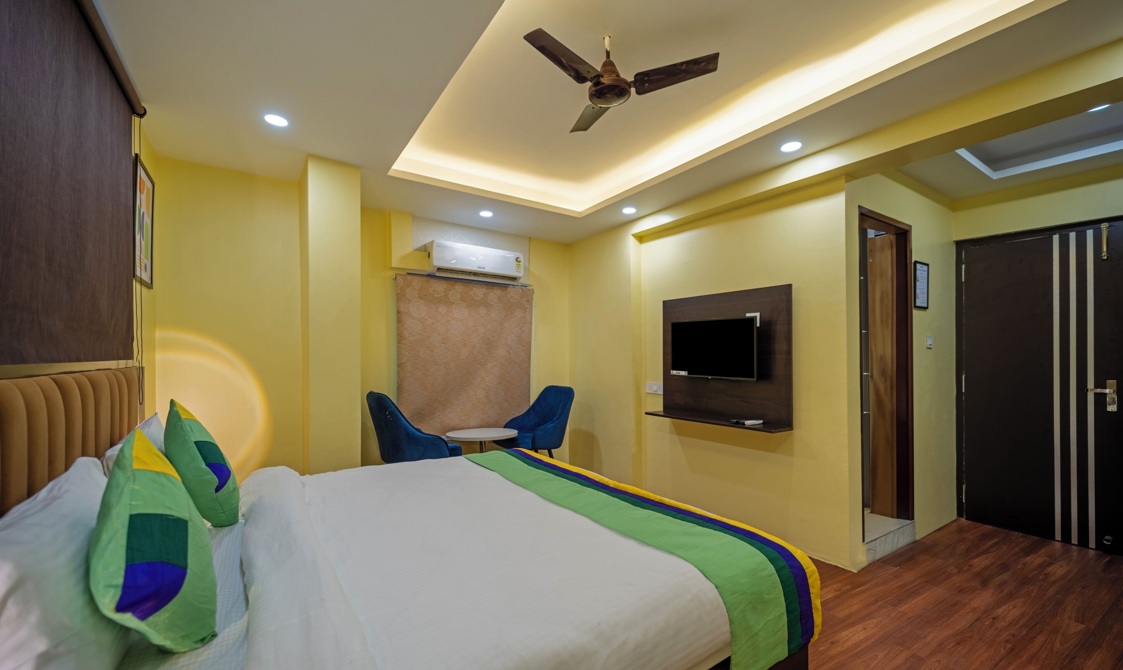 A hotel room with a bed, a ceiling fan, an air conditioning unit, a TV mounted on the wall, and a door.