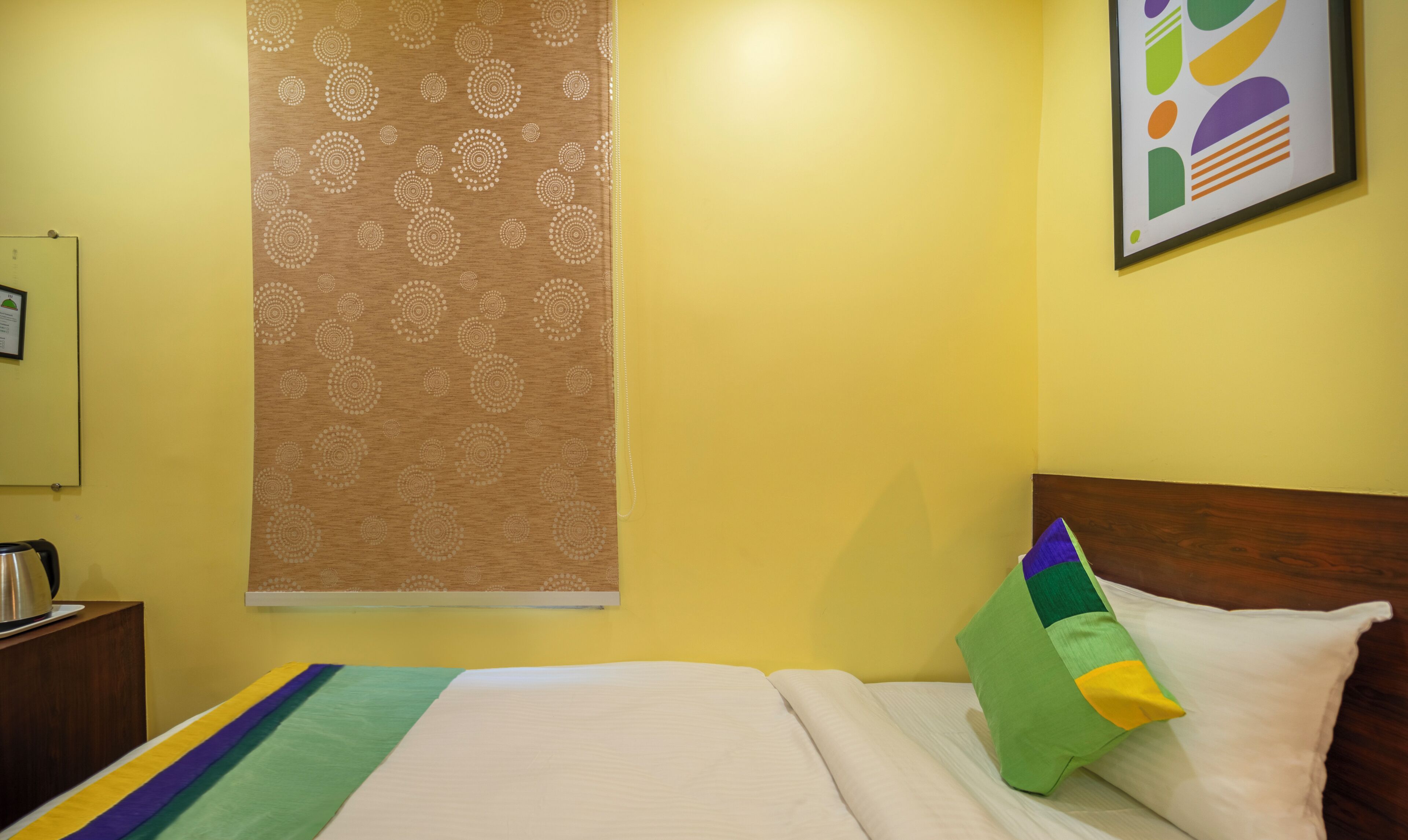 Een hotelkamer met een bed, nachtkastje, waterkoker, een kamerbehang met patroon en een kleurrijk abstract schilderij.