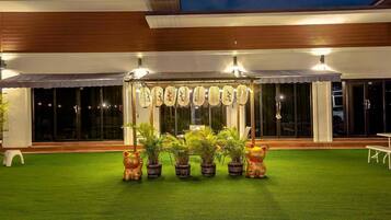Un área de estar al aire libre con techo, césped artificial, plantas en macetas y un toldo decorativo.