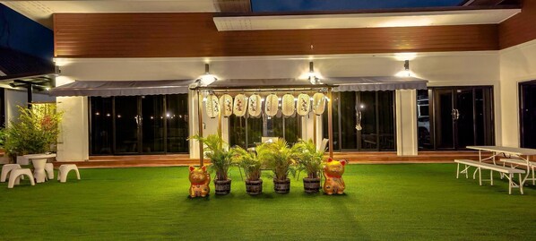 Un área de estar al aire libre con techo, césped artificial, plantas en macetas y un toldo decorativo.