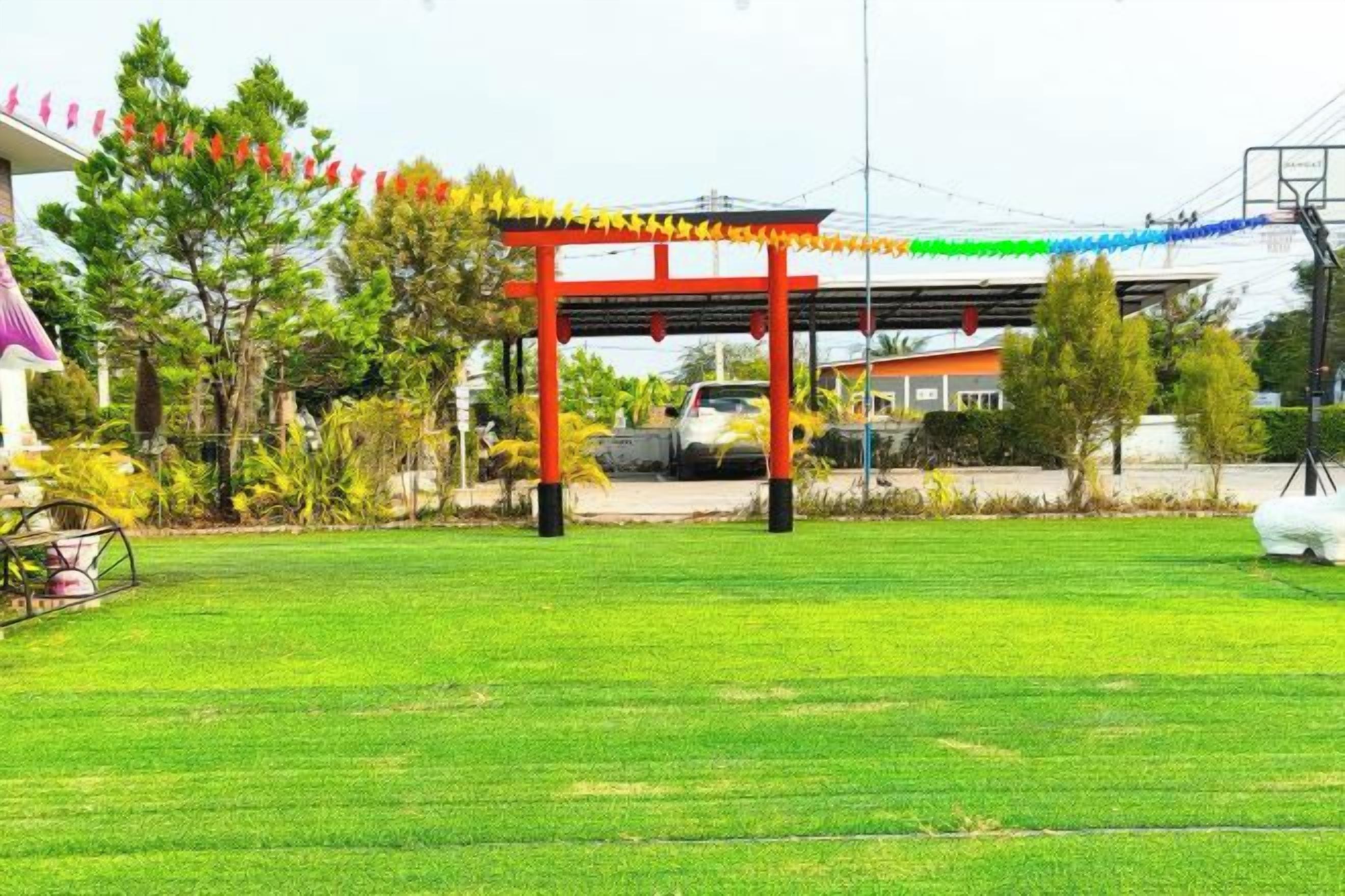 A red torii gate with a green and yellow rope decoration, a grassy field, and a building with a pink roof in the background.