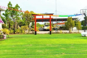 A red torii gate with a green and yellow rope decoration, a grassy field, and a building with a pink roof in the background. - The Chill Resort Resort at Korat (Nakhon Ratchasima)