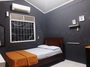 A small, dark-colored room with a single bed, a window with a metal grid, a wall-mounted air conditioner, and a small shelf with a picture and a phone. - Hotel O Omah Citra Genia Homestay (Ngaglik)