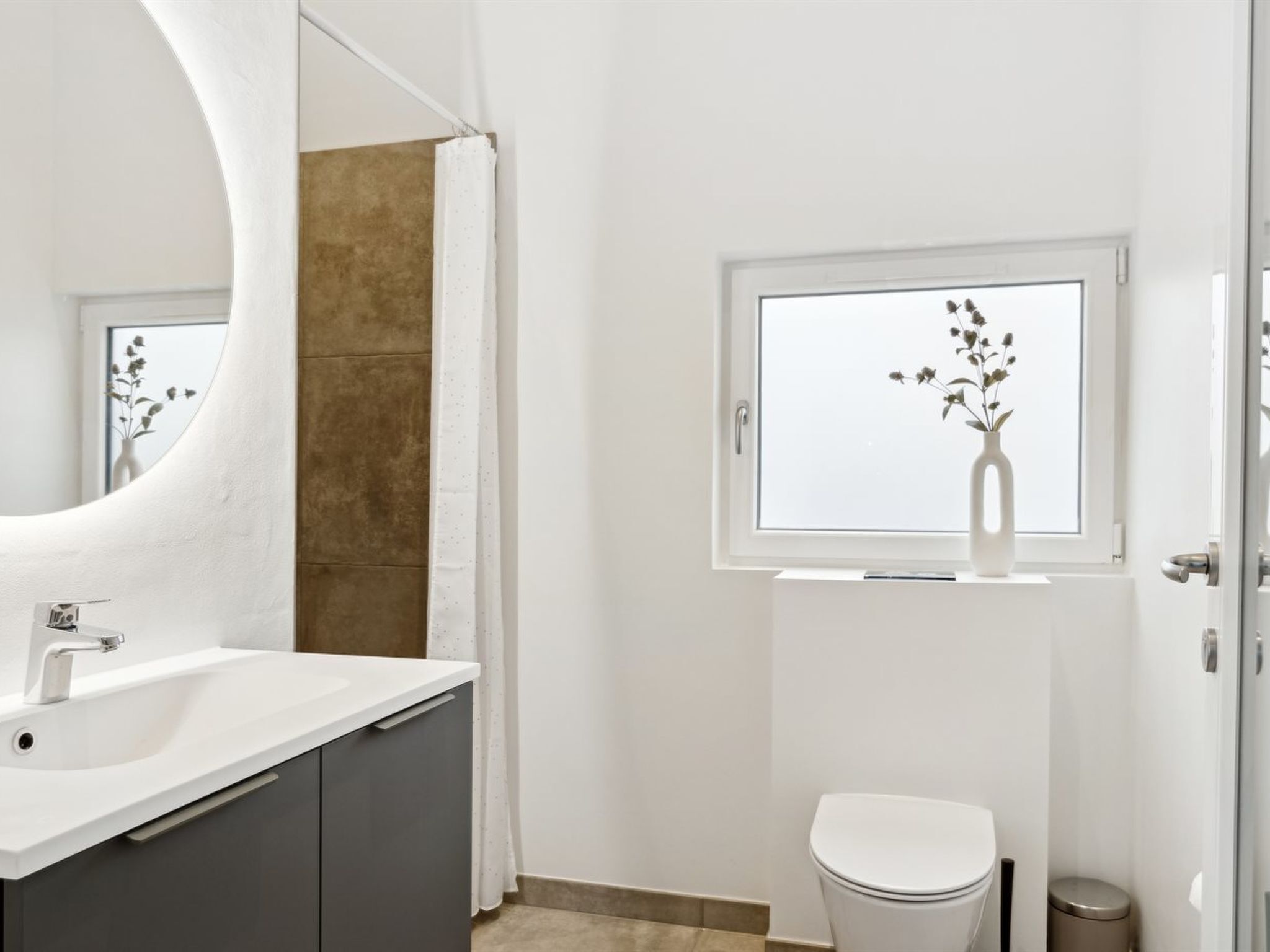 Un bagno moderno con un lavandino bianco, uno specchio grande, una finestra vista fiori e un WC.