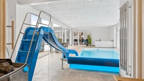 Une piscine intérieure moderne dotée d’un toboggan bleu, d’un espace clos par des parois de verre et d’un coin salon bien visible, offrant une vue sur la piscine.
