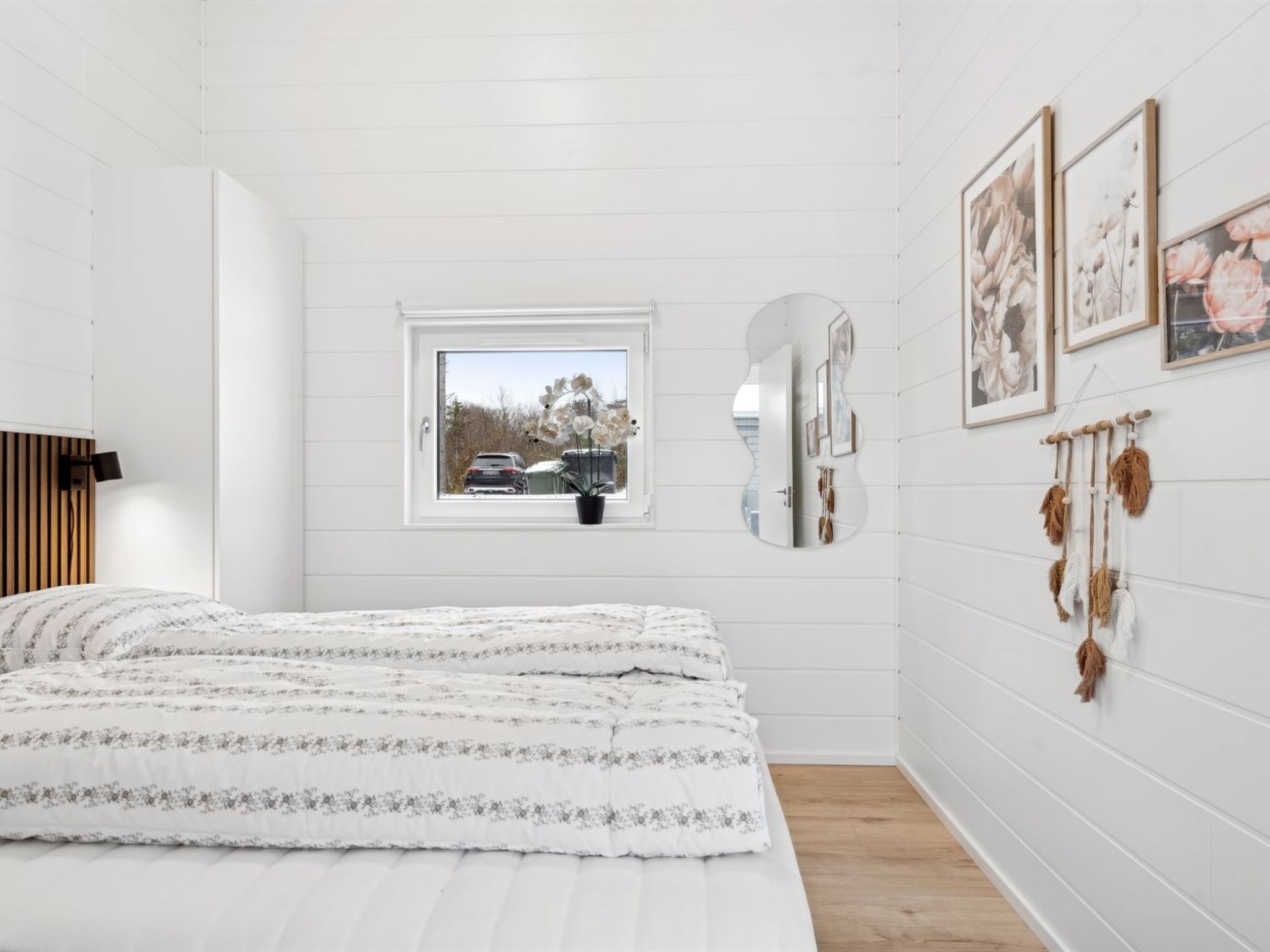 A bedroom with a bed, a window with a view of parked cars, a mirror, and wall-mounted decorations.