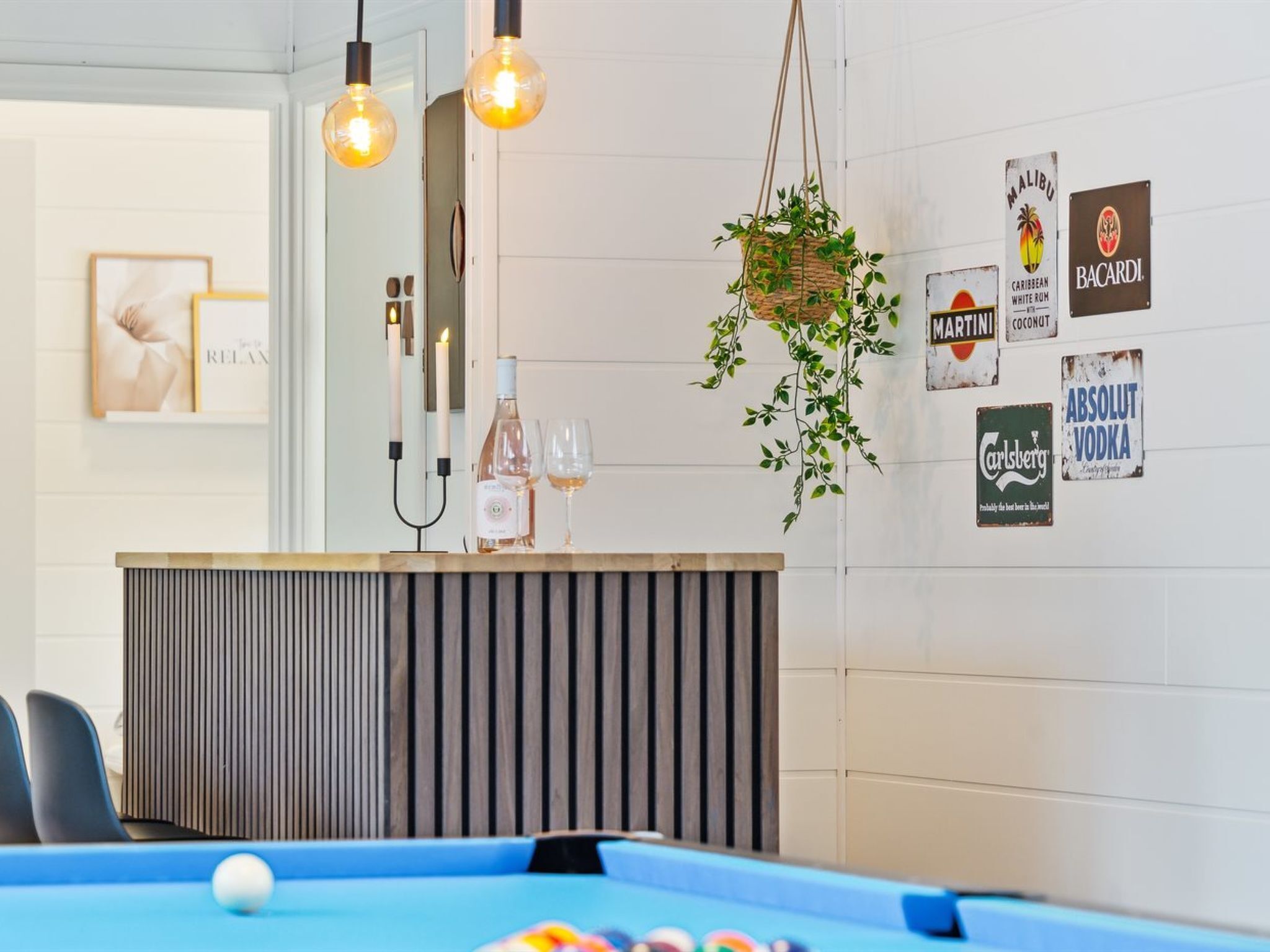 Une table de billard avec une boule blanche, une bouteille de vin et des verres posés sur un comptoir ; une plante suspendue ; et des éléments de décoration murale, notamment des logos de marques.
