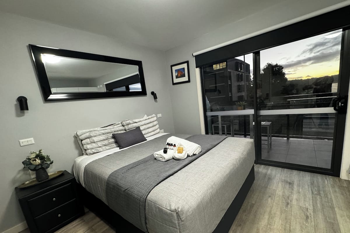 Una camera da letto moderna con un letto grande, un comodino, uno specchio e una porta scorrevole in vetro che dà su un balcone.