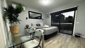 Ein modernes Schlafzimmer mit einem Glastisch, einem Bett mit grauer Bettwäsche, einer Topfpflanze und einer Glasschiebetür, die zu einem Außenbereich führt.