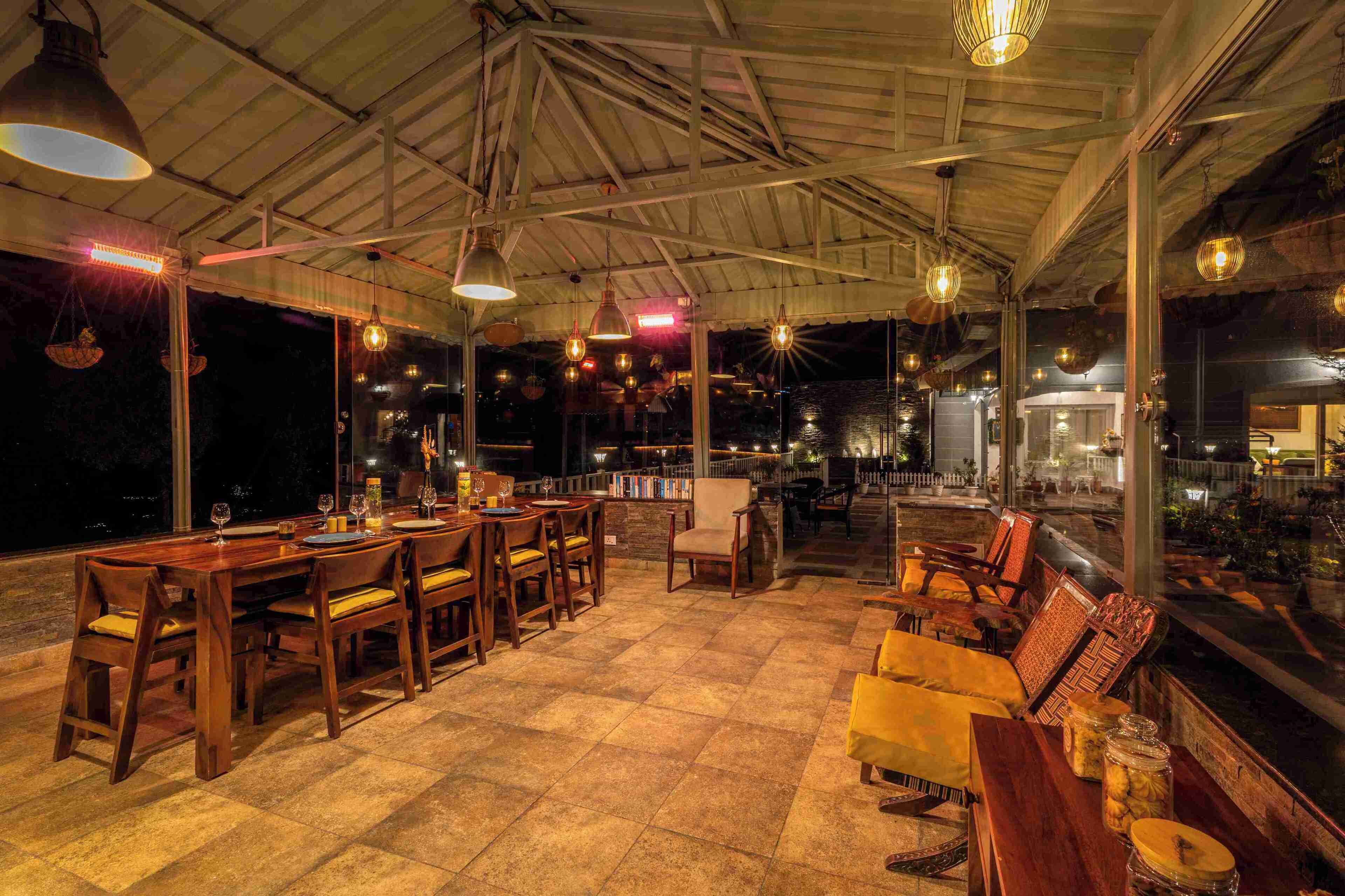 An outdoor dining area with wooden tables and chairs, large hanging lights, and a view of the water.