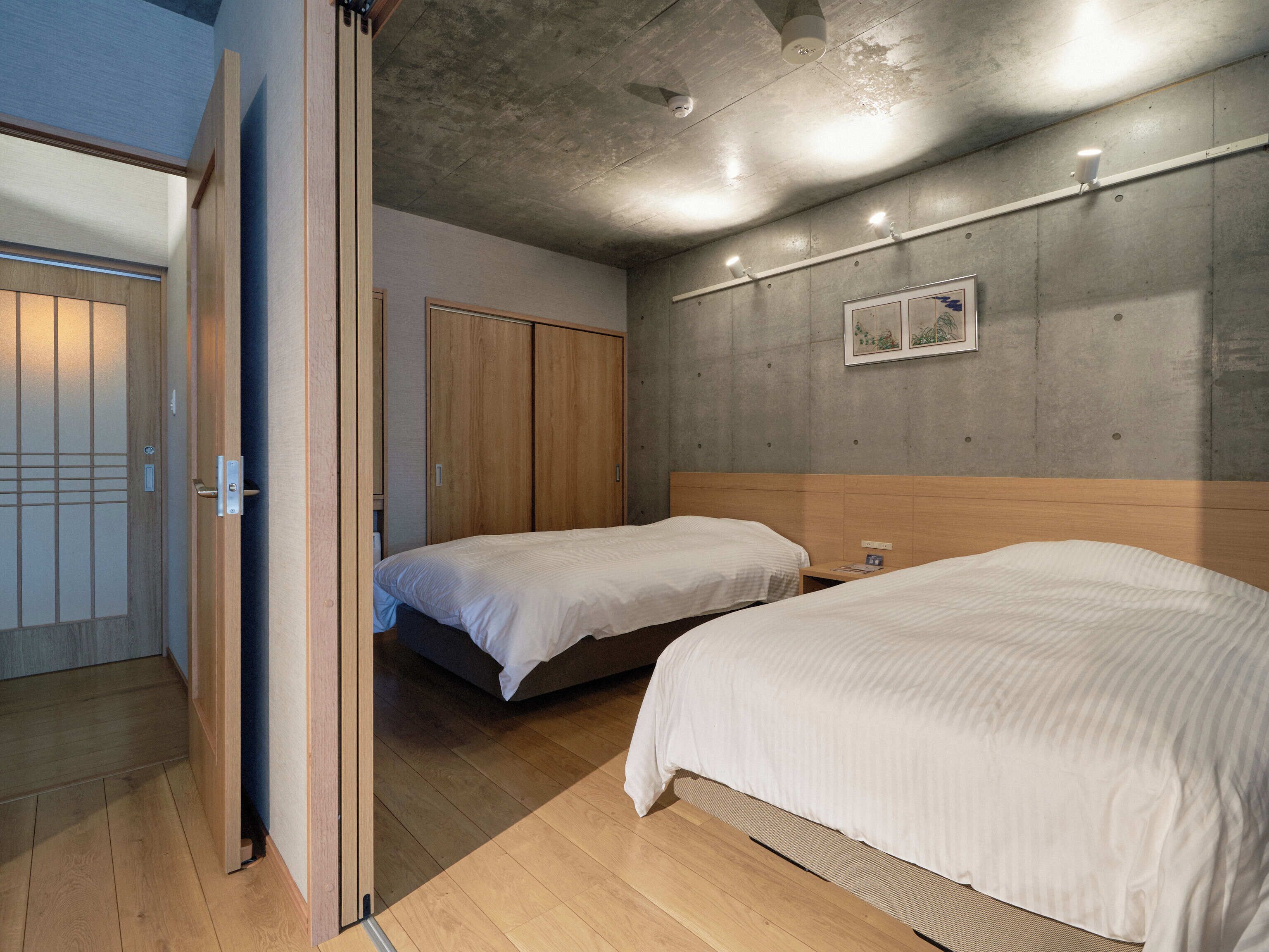 Ein Hotelzimmer mit zwei Einzelbetten, Holzboden, einer Betonwand als Gestaltungselement und einer Tür mit traditionellem Design.