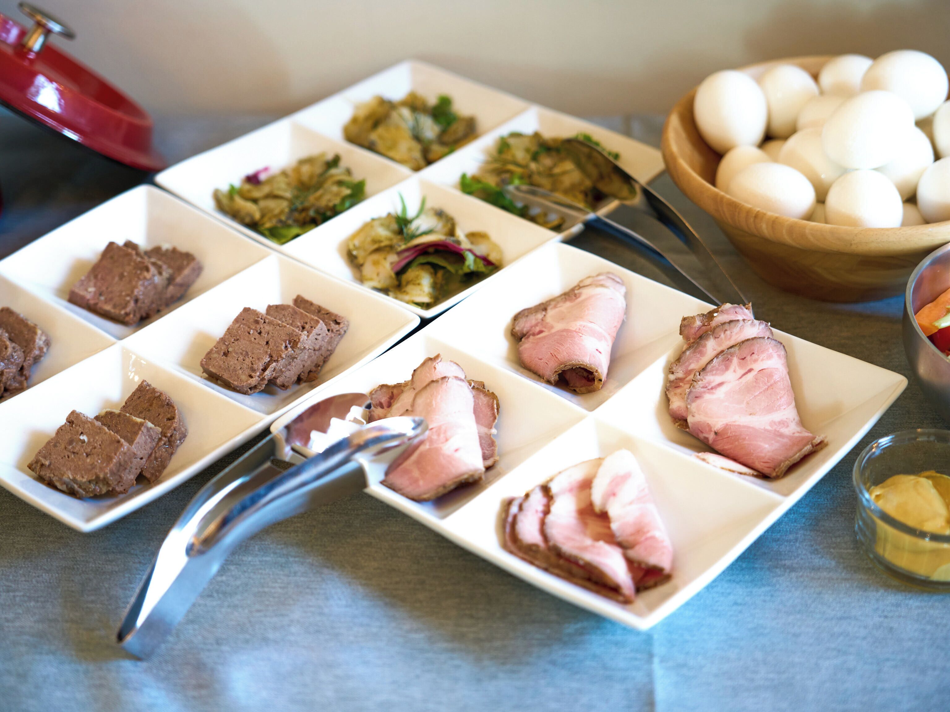 A table with various food items including meat, vegetables, and eggs, with a pair of tongs on it.