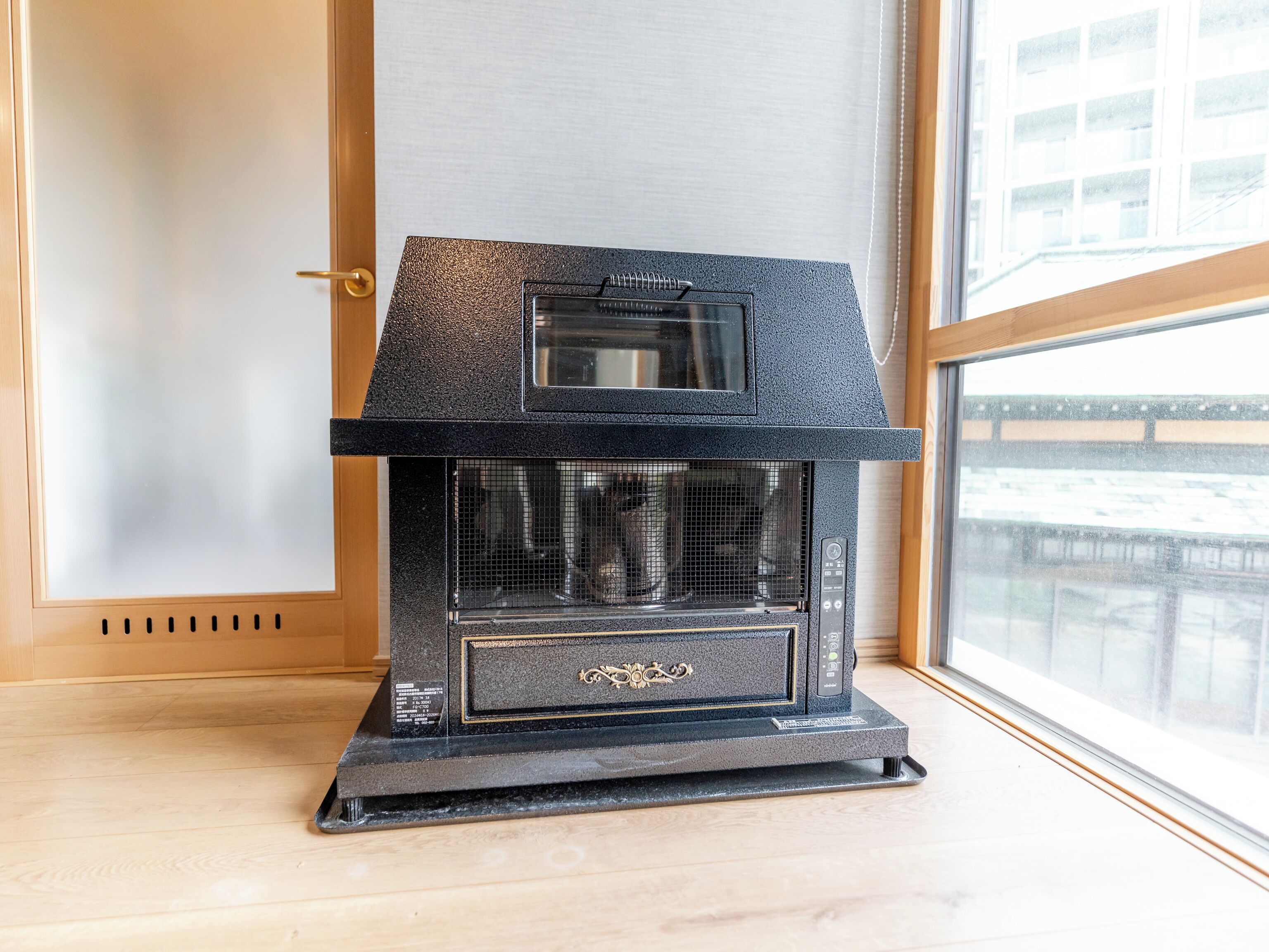 A black wood stove with a glass window and a control panel, placed in a room with a wooden floor and a window with a view of buildings.