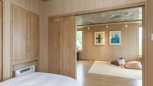 Ein Schlafzimmer mit Bett, Holzschrank und Blick auf eine Yogamatte und Kissen im angrenzenden Raum.
