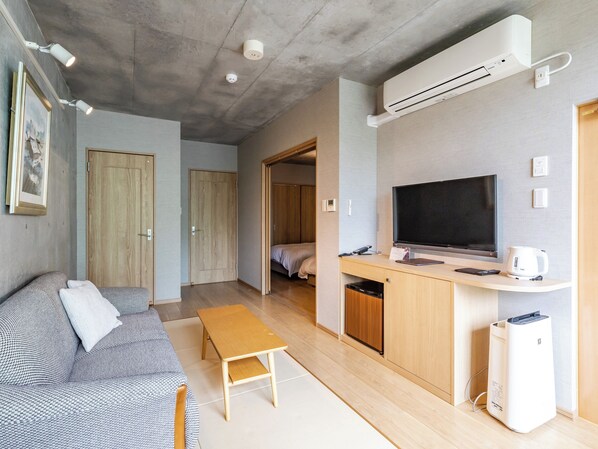 Un espacio habitable compacto con sofá, una pequeña mesa de madera, una kitchenette y un televisor, además de aire acondicionado instalado en el techo.
