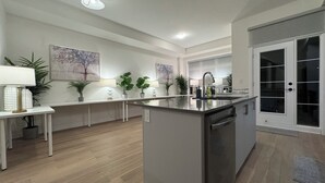 A modern kitchen with a central island, white cabinetry, and a built-in dishwasher. There are several potted plants and a painting on the wall. - Elegant Townhome in Prestigious Woodbridge Estate, Vaughan (Selena) (Vaughan)