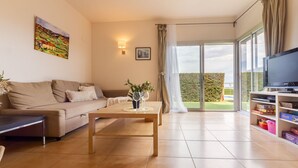 Una sala de estar con sofá, mesa de centro y televisor sobre un soporte. Hay un ventanal con cortinas y vistas al exterior con vegetación.