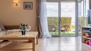 Una sala de estar moderna con un sofá, una mesita de centro con bebidas, un televisor y un ventanal corredizo a un balcón con sillas y plantas.