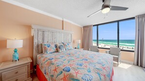 Een slaapkamer met een groot bed, een plafondventilator, een nachtkastje met lamp, uitzicht op het strand en een raam met gordijnen.