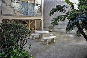 Een binnenplaats met een stenen bank en tafel, omgeven door potplanten en een gebouw met een balkon.