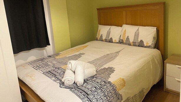 Een netjes opgemaakt bed met een bedspreide met patroon, twee kussens met een stadsgezicht dessin, een houten hoofdbord en een klein nachtkastje.