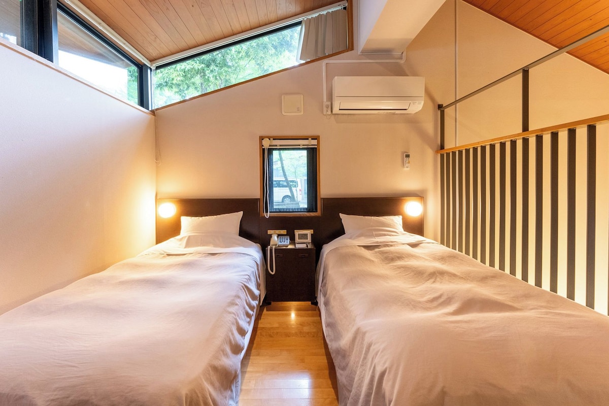A hotel room with two beds, a window with a view of a vehicle, an air conditioning unit, and a bedside telephone.