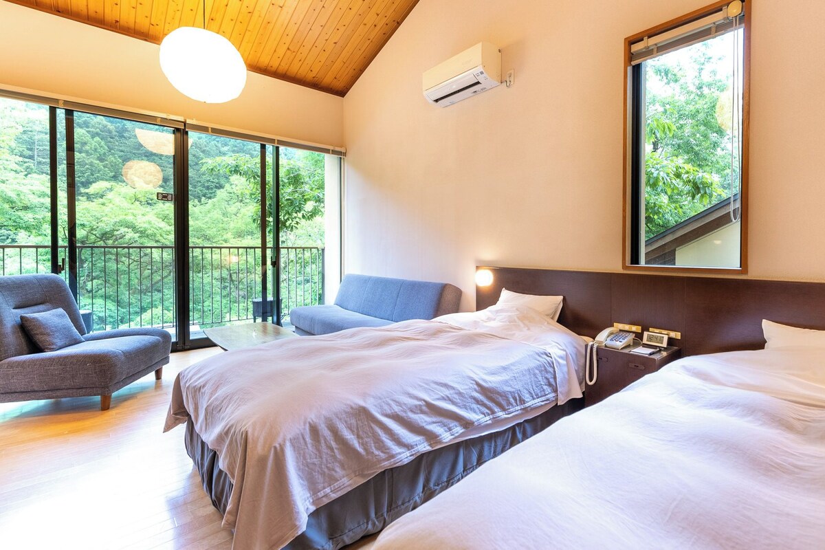 A hotel room with two beds, a sofa, a balcony with a view of greenery, and a large window.