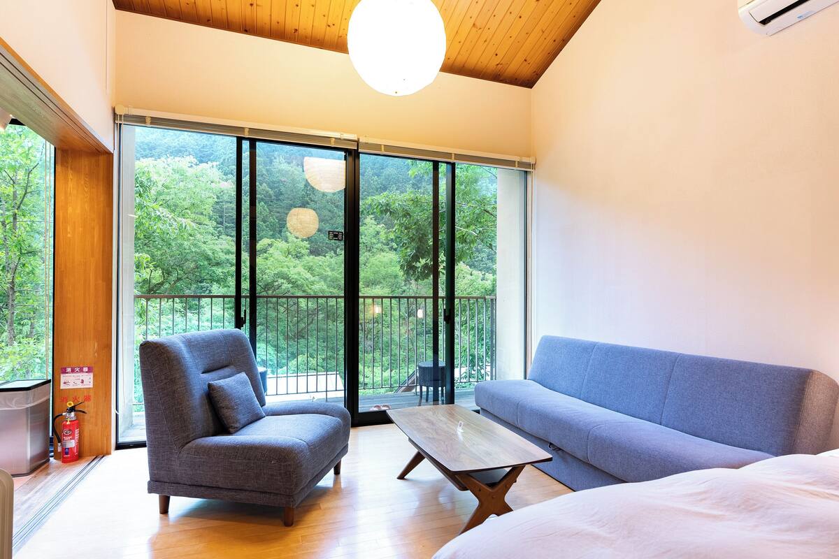 A modern room with a wooden ceiling, a large sliding glass door leading to a balcony, a grey sofa, a grey armchair, and a wooden coffee table.