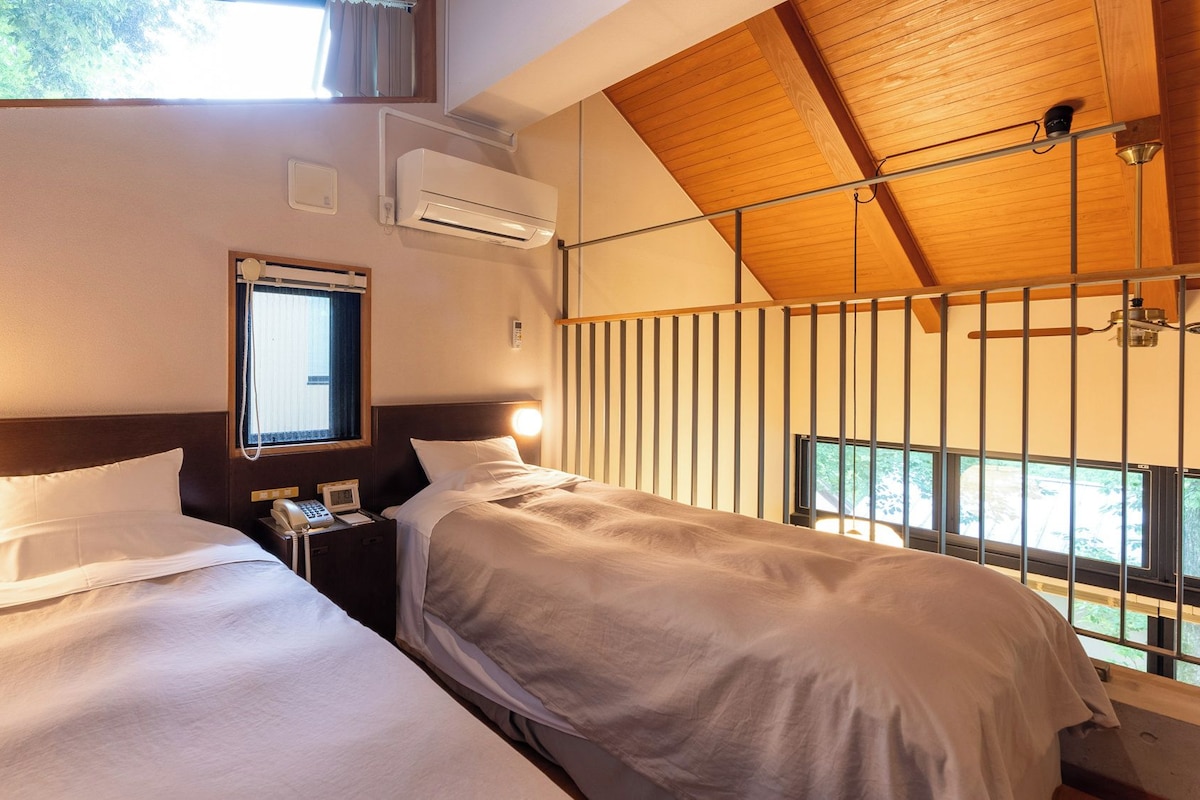 A hotel room with two beds, an air conditioning unit, a bedside telephone, and a window with a view.