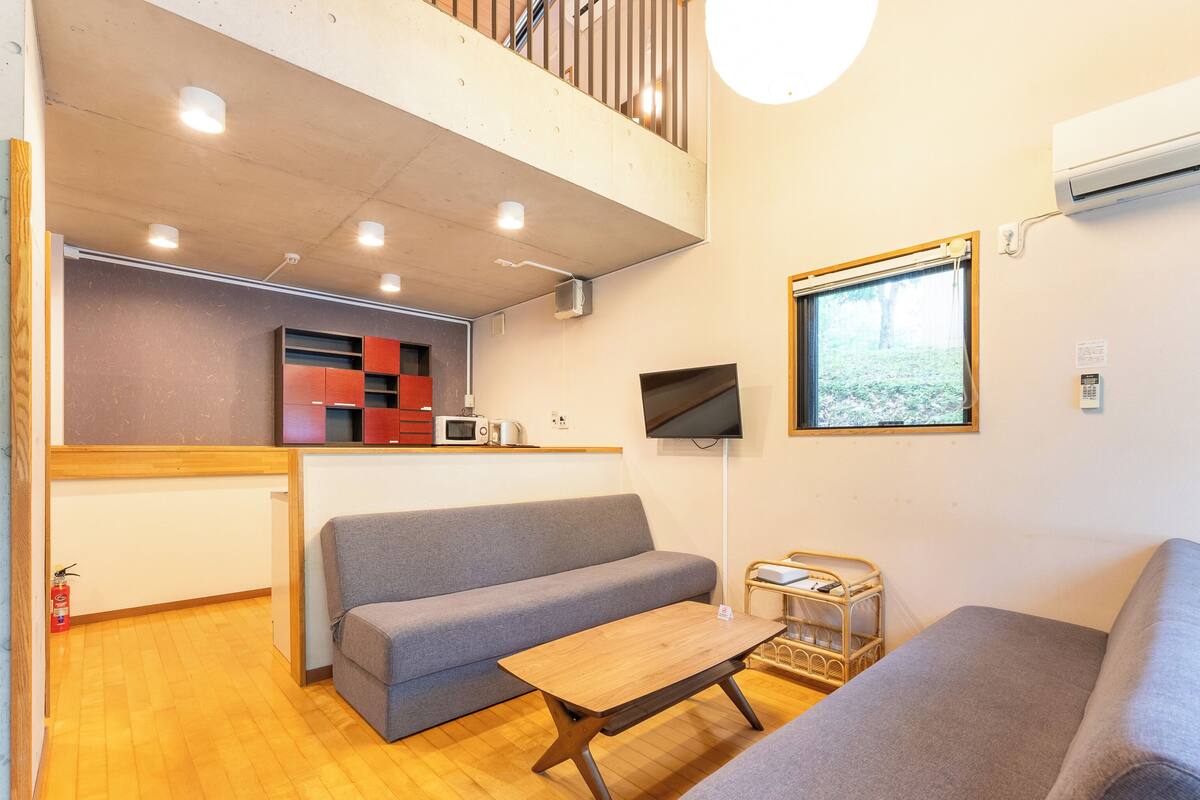 A compact living space with a kitchenette, a grey sofa, a wooden table, and a TV mounted on the wall.