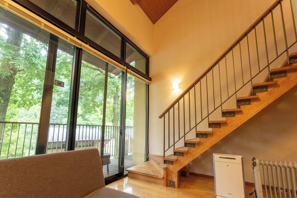 A modern interior with a wooden staircase, a sofa, and a view of greenery through large windows.