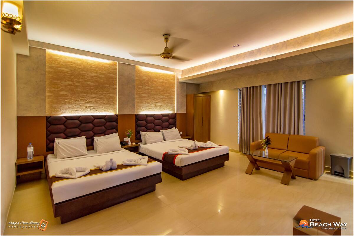 A hotel room with two beds, a sofa, a coffee table, and a ceiling fan.