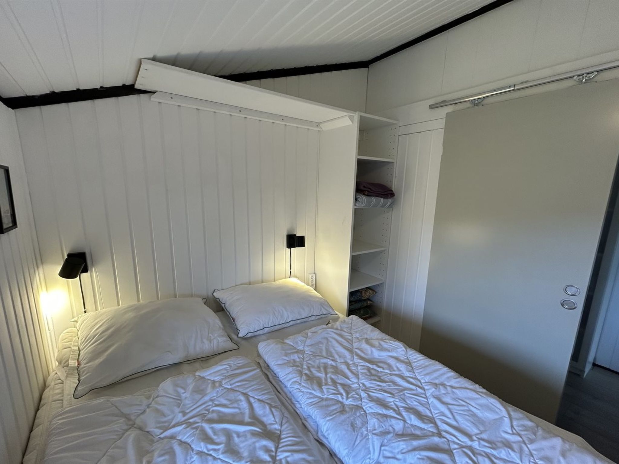 Una cama individual con ropa blanca, dos mesitas de noche fijadas a la pared, un armario y una estantería con objetos doblados.