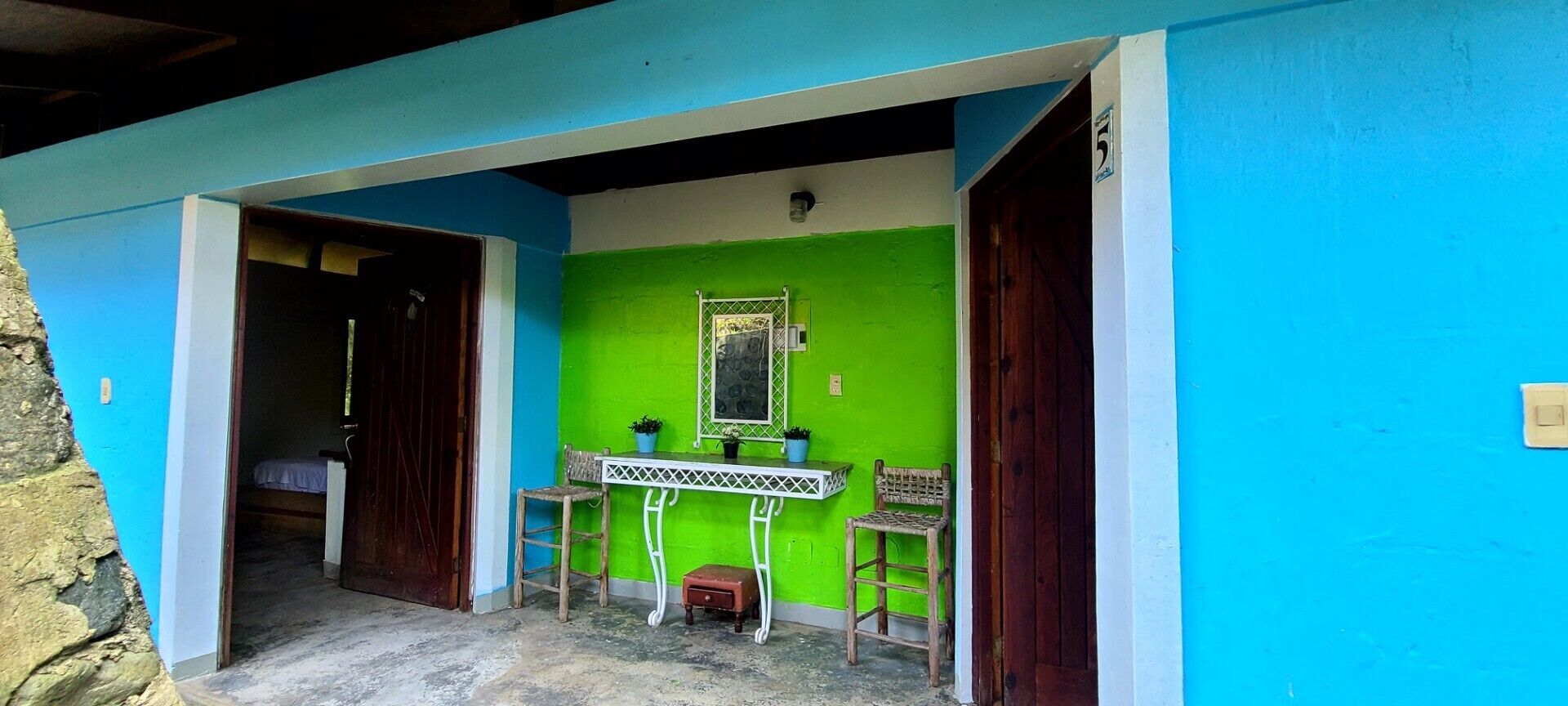 Una habitación con una pared verde brillante, una mesa blanca con dos sillas, un espejo pequeño y una puerta que da a otra habitación.