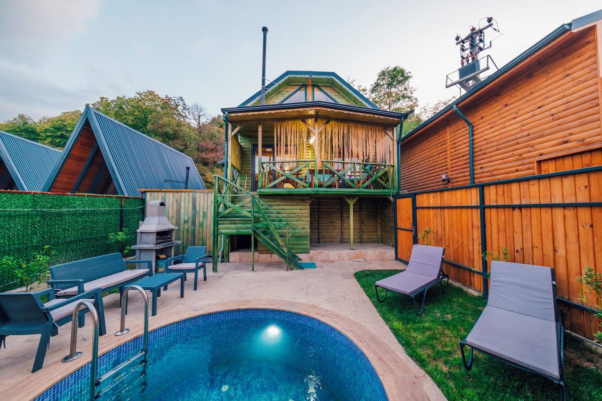 A backyard with a swimming pool, outdoor furniture, a barbecue grill, and a wooden cabin structure.