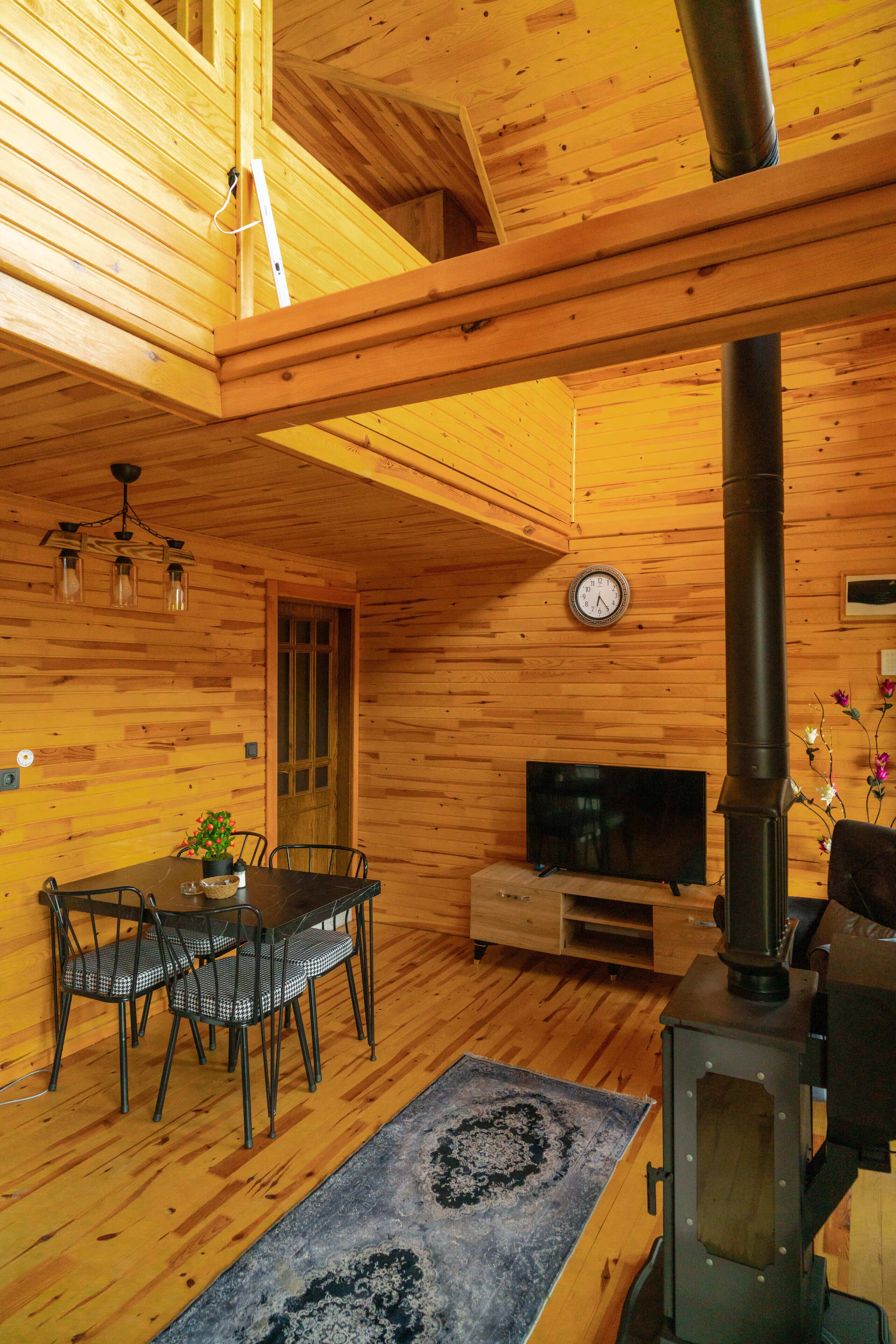 A wooden cabin interior with a dining area, a TV, a clock, and a wood-burning stove.