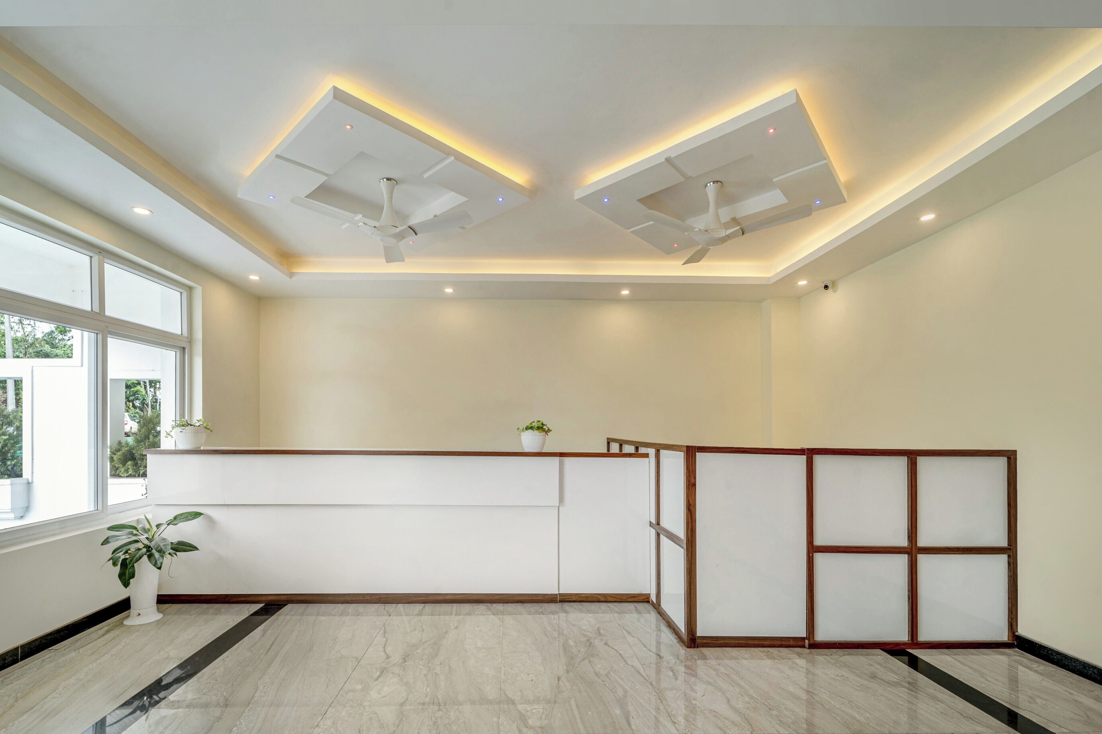 A hotel reception area with a modern design, featuring a reception desk, two ceiling fans, and a large window providing natural light.